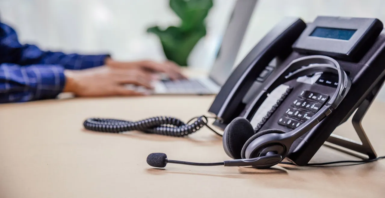 Desk phone with attached headset and person using a laptop in the background. Phone systems in Halfmoon NY