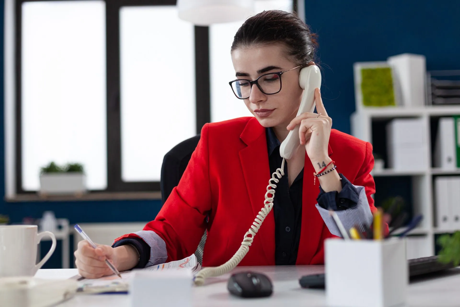 Woman in red blazer on landline phone, writing at a desk in an office. Support services in Halfmoon